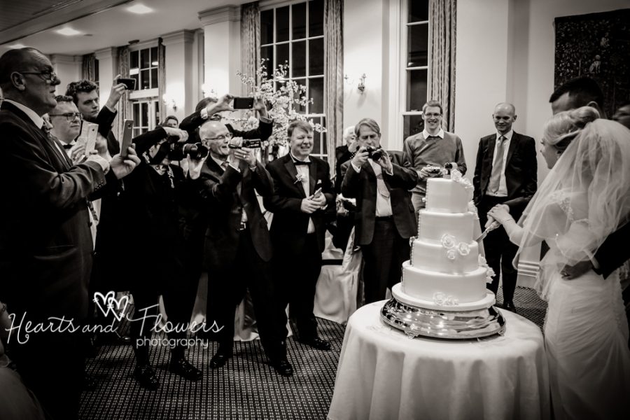 A crowd of guests taking pictures of the bride and groom cutting the cake.