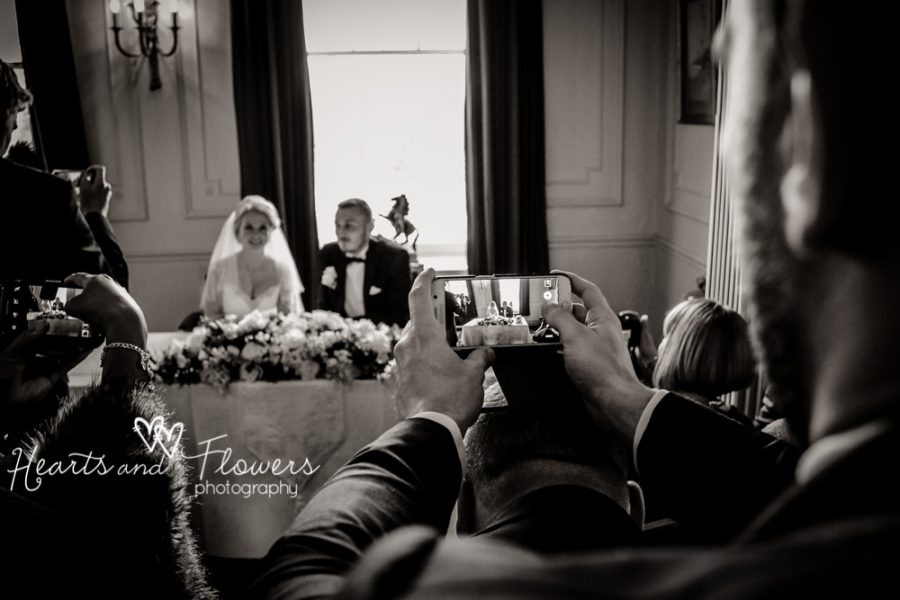 Cameras are taking pictures of the bride and groom sitting at the table with the flowers.